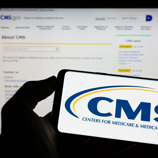 A hand holding a phone displaying the CMS logo in front of a computer screen displaying its website -- Insurance coverage from STAT